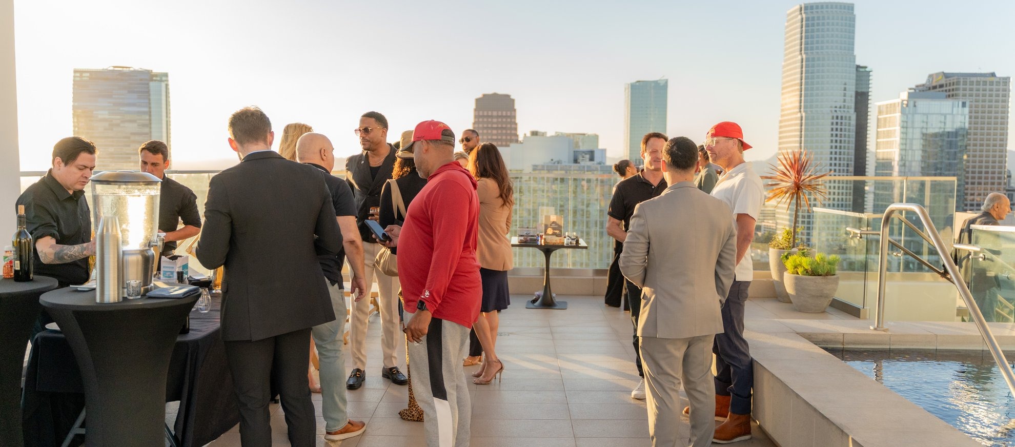 A group of people in semi-formal attire socialize on a rooftop terrace with city skyscrapers in the background and a pool nearby, under bright sunlight.