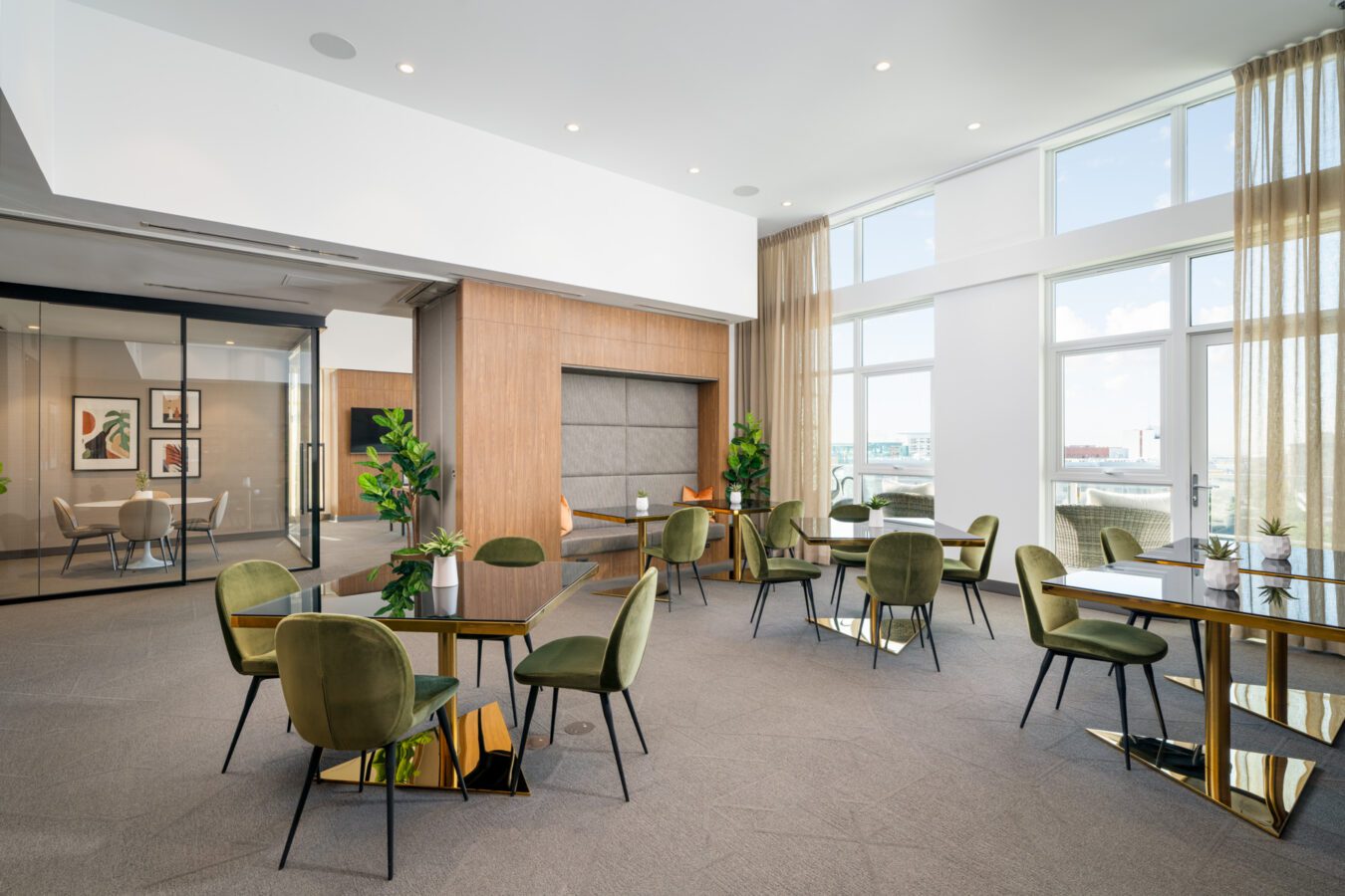 A modern, spacious office lounge with glass tables, green velvet chairs, potted plants, large windows with sheer curtains, natural light, and framed artwork on the walls.