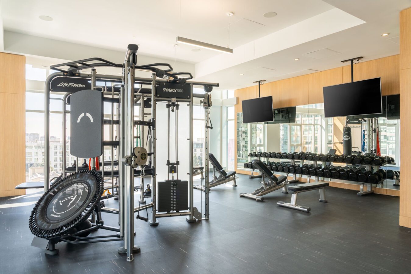 A modern gym with large windows, strength training equipment, a set of dumbbells with benches, a rebounder, two wall-mounted TVs, and mirrors reflecting the bright, spacious room.