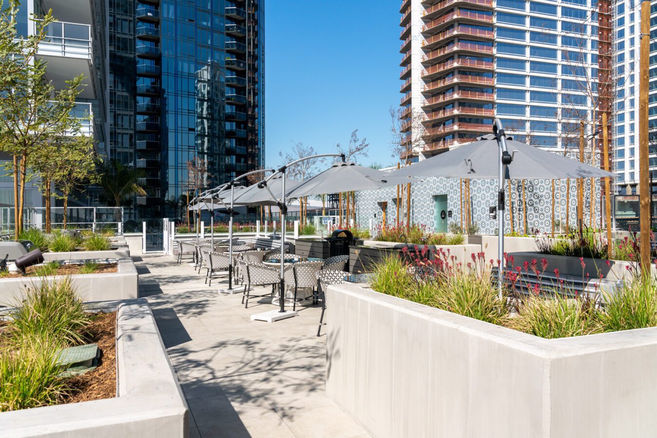 Modern outdoor patio with tables, chairs, and gray umbrellas, surrounded by planters with greenery. Tall glass and brick high-rise buildings are visible in the background under a clear blue sky.