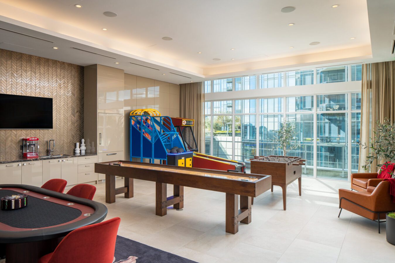 A modern game room with a poker table, shuffleboard, foosball table, and basketball arcade game; large windows let in natural light, and there are red chairs, a TV, and a coffee maker on the counter.