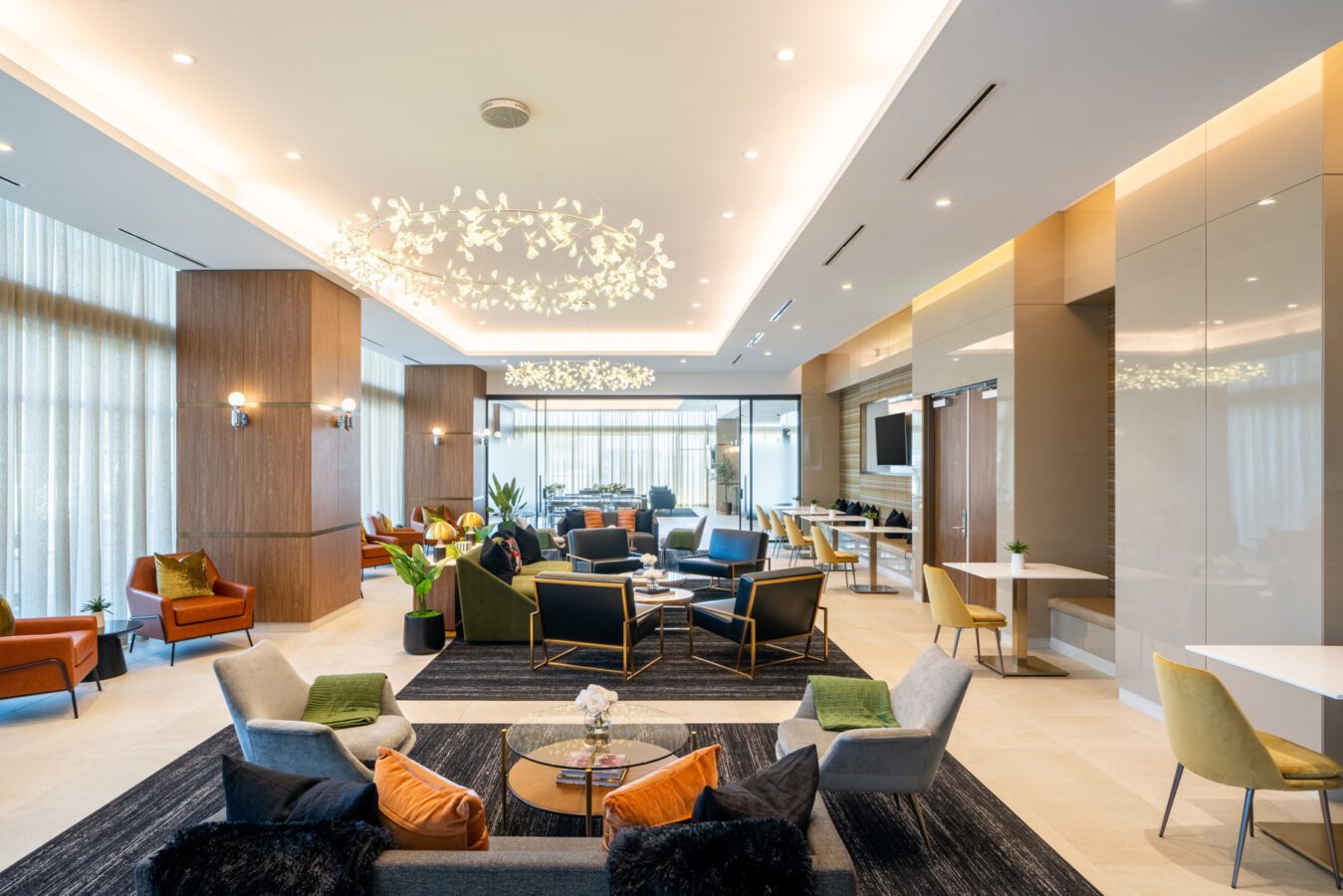 A modern, spacious lounge with contemporary furniture, soft lighting, and floor-to-ceiling windows. Armchairs and sofas are arranged around coffee tables, with decorative lighting hanging from the ceiling.