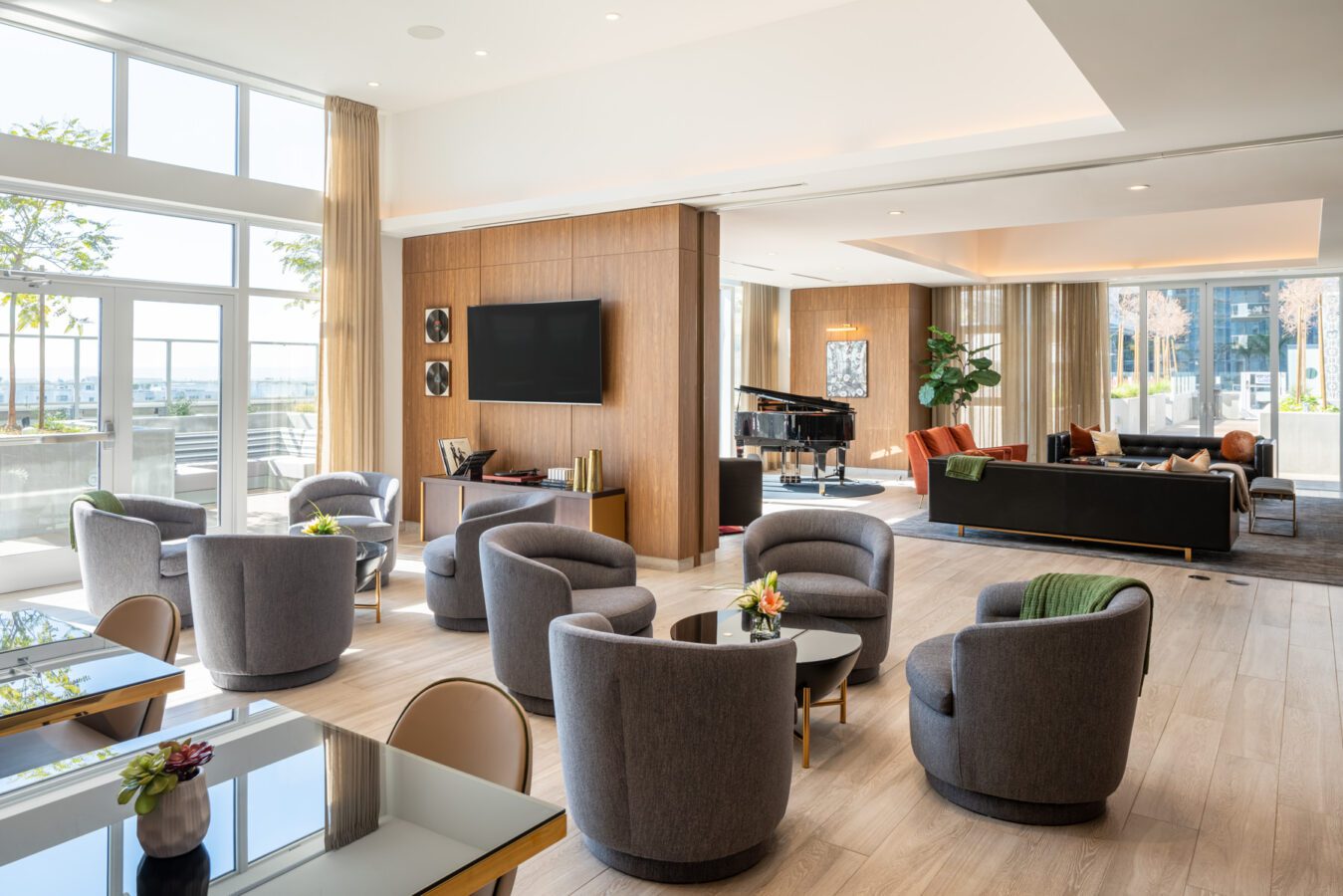 Modern, spacious lounge with large windows, gray armchairs, round tables with small floral arrangements, a TV on a wood-paneled wall, a grand piano, and a black sofa in the background. Natural light fills the room.
