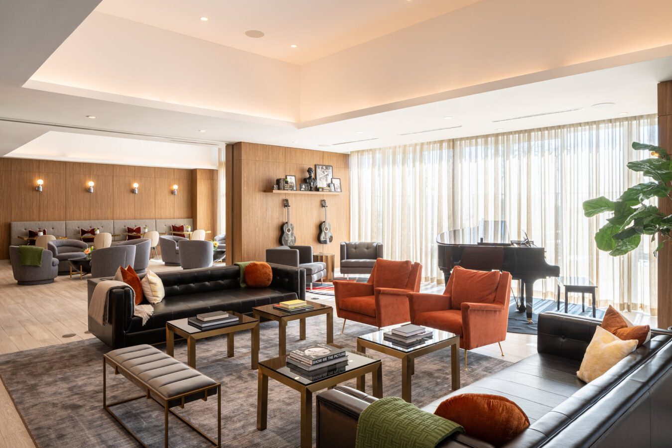 Modern lounge with black leather sofas, orange chairs, a grand piano, and coffee tables on a large rug. Floor-to-ceiling sheer curtains let in natural light. Shelves, plants, and guitars decorate the wood-paneled walls.