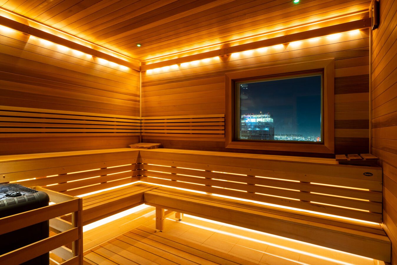 A modern wooden sauna with warm, ambient lighting, built-in benches, and a window overlooking a cityscape at night.