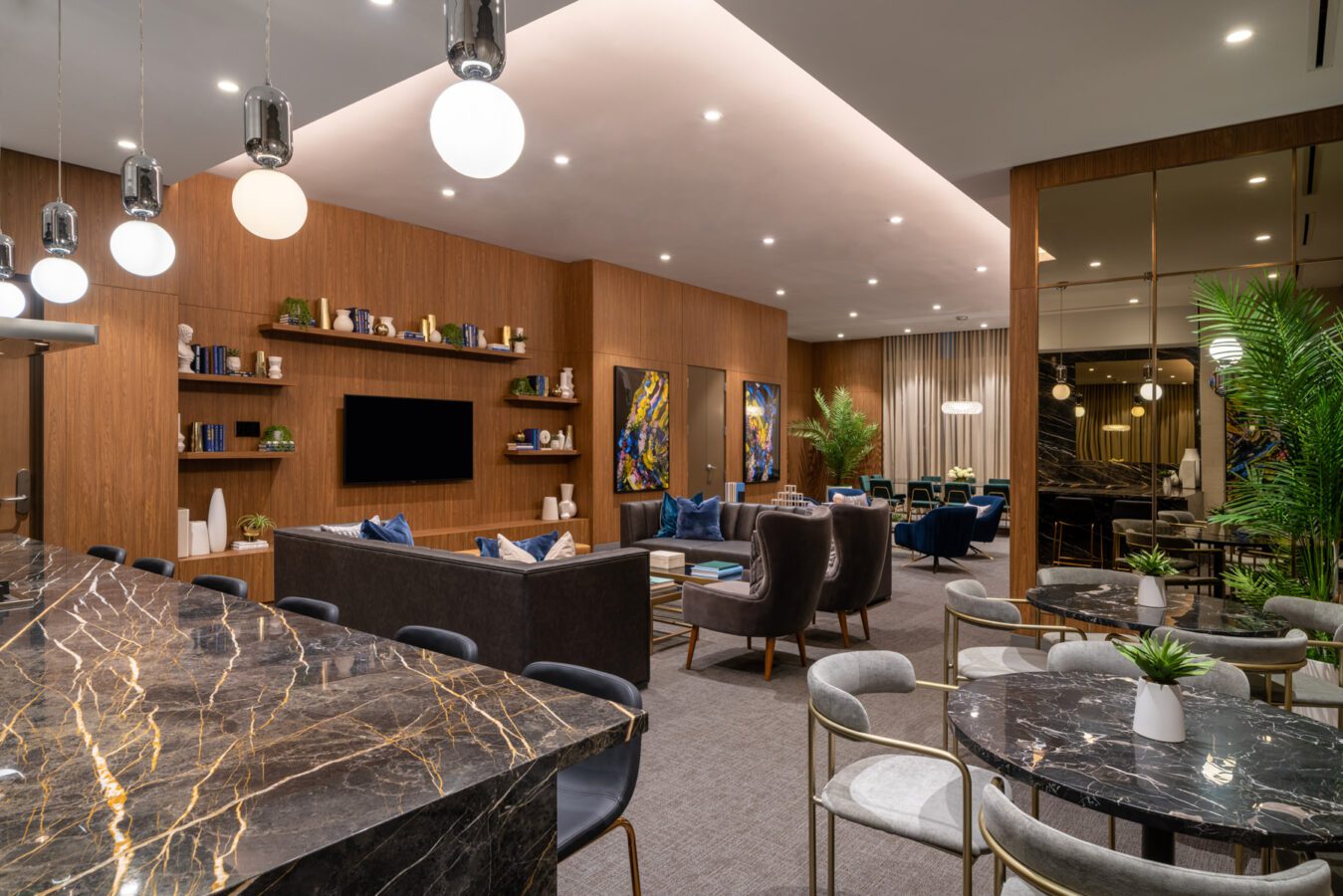 Modern lounge with marble tables, sofas, armchairs, and decorative plants. Wooden panel walls feature shelves, art, and a TV. Pendant lights and recessed ceiling lights illuminate the stylish, inviting space.