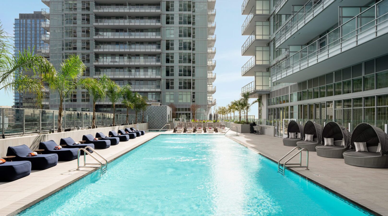 Modern outdoor swimming pool surrounded by lounge chairs and palm trees, situated between two tall glass apartment buildings under a clear blue sky.
