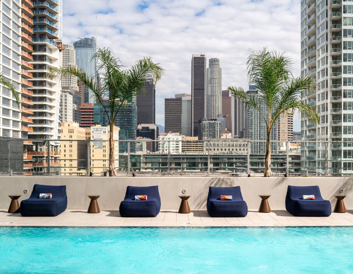 A rooftop pool with turquoise water, lined with navy blue lounge chairs and small tables, overlooks a city skyline with tall buildings and palm trees under a partly cloudy sky.