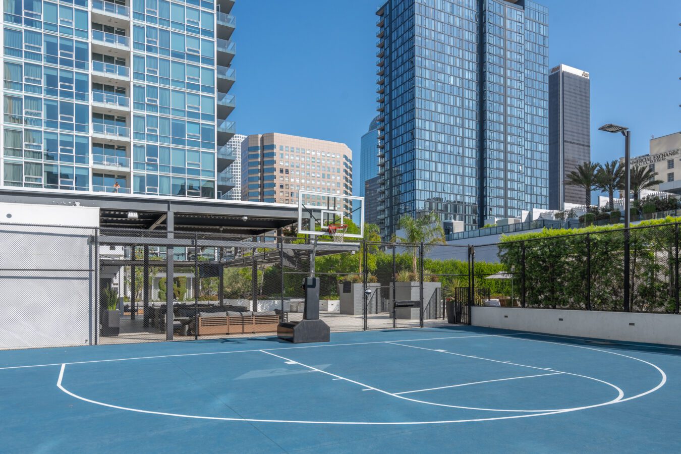 Outdoor blue basketball court with white lines, surrounded by a wire fence and greenery, located in an urban area with modern high-rise buildings in the background under a clear blue sky.