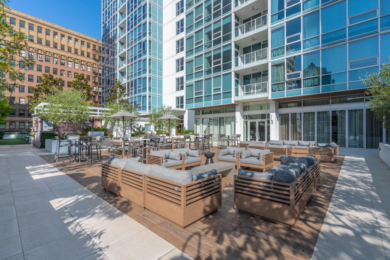 Modern outdoor patio area with wooden seating, tables, and chairs, surrounded by plants, adjacent to a tall glass apartment building in an urban setting on a sunny day.