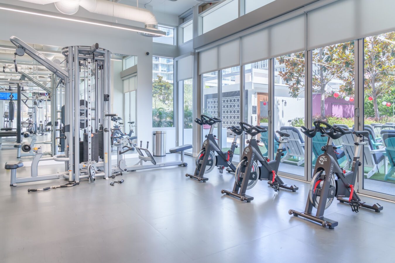 Bright gym with large windows, three stationary bikes, weight machines, and exercise equipment. Outside, there is patio seating with umbrellas and greenery visible through the glass.