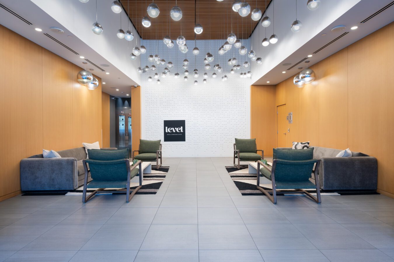 Modern lobby with light wood walls, gray tile floor, green and gray lounge chairs, sofas, and hanging glass globe lights. A white wall at the back displays the “level” logo in black.