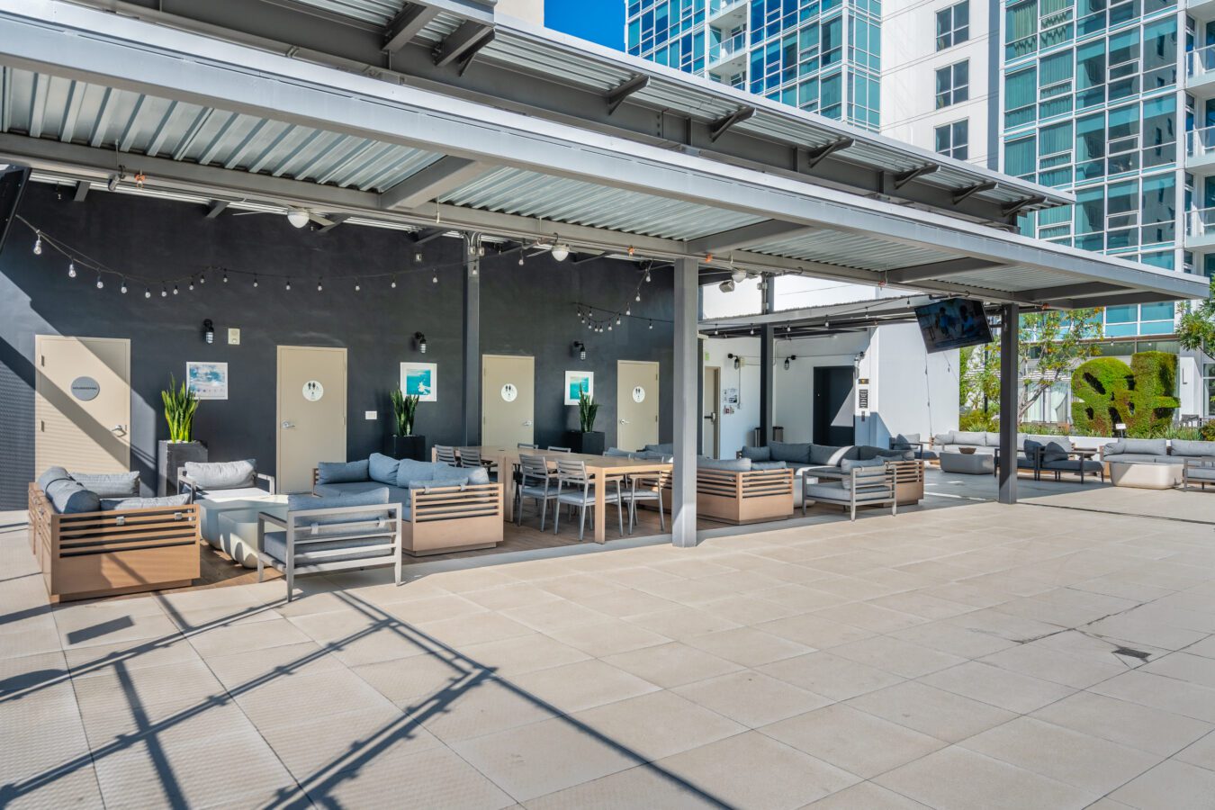 Modern outdoor lounge area with wooden and gray cushioned seating, tables, and potted plants under a metal roof. High-rise buildings and string lights are visible in the background.