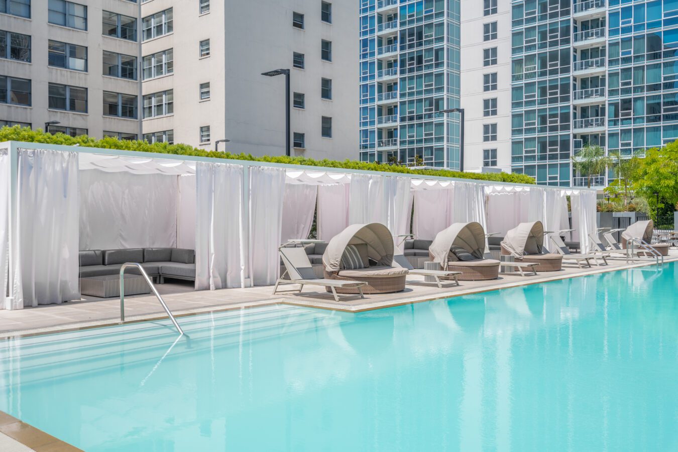 A modern outdoor pool with clear blue water, lined with lounge chairs and shaded cabanas draped in white curtains, surrounded by tall glass and concrete buildings.