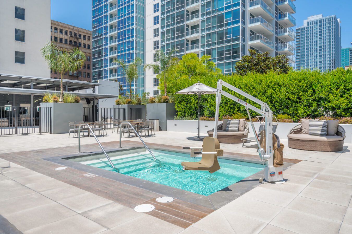 Outdoor hot tub on a modern apartment patio with lounge chairs, umbrella, greenery, and a pool lift for accessibility. Tall glass buildings and palm trees are visible in the background.