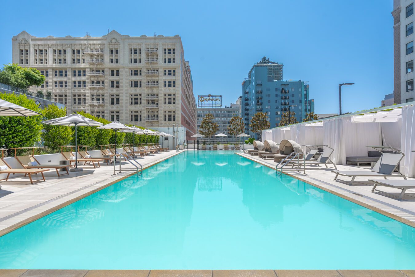 A rooftop swimming pool with lounge chairs, sun umbrellas, and cabanas on both sides. Tall buildings and clear blue sky are visible in the background. The scene is bright and modern.
