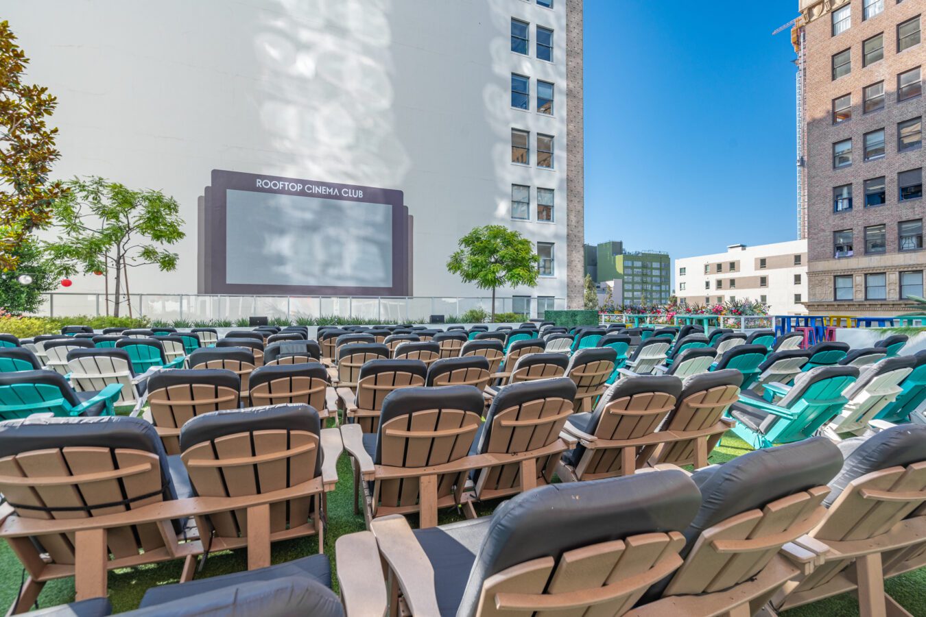 Rows of empty outdoor chairs face a large movie screen at a rooftop cinema, surrounded by tall buildings and greenery under a clear blue sky.