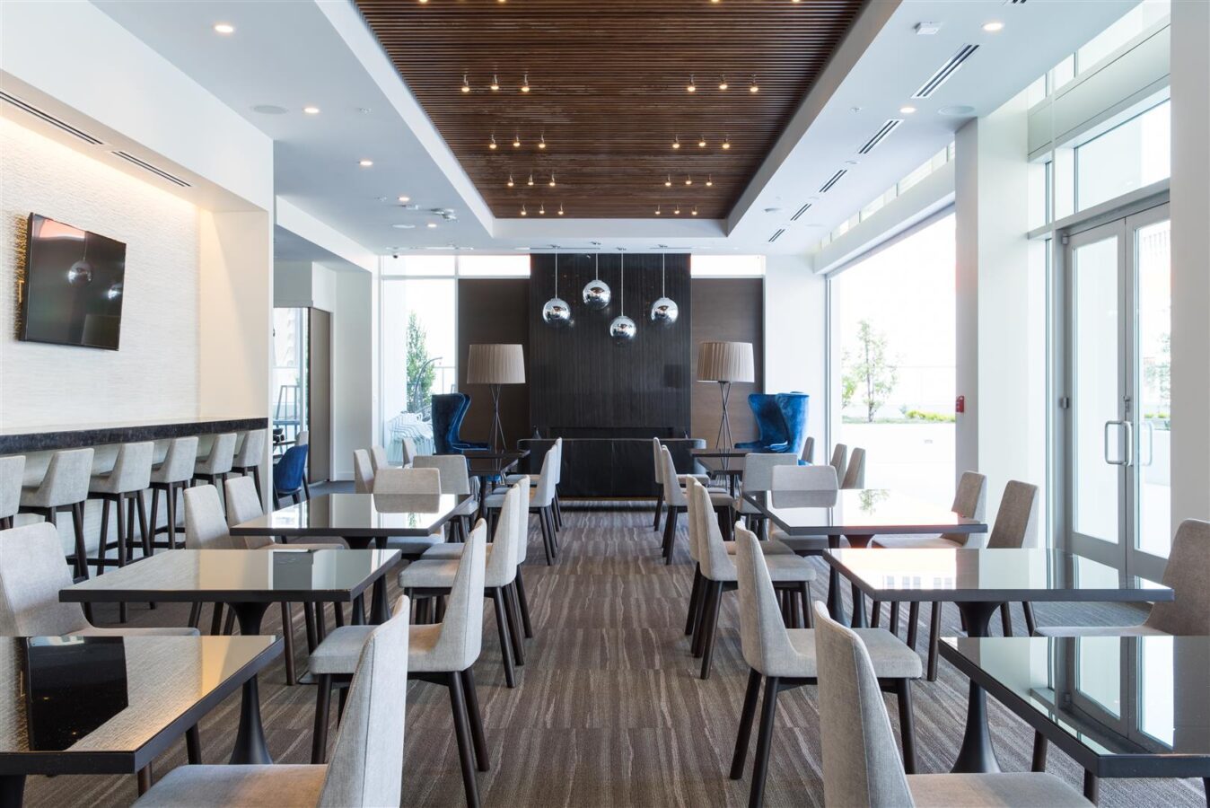 Modern dining area with neatly arranged black tables and beige chairs, pendant lights above, large windows letting in natural light, and minimalist decor with some blue accent chairs in the background.
