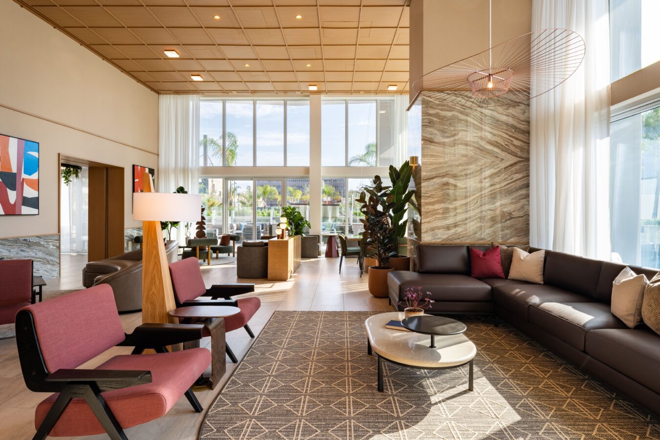 A modern hotel lobby with large windows, a sectional sofa, pink and black chairs, a geometric rug, tall plants, and abstract wall art, filled with natural light and contemporary decor.