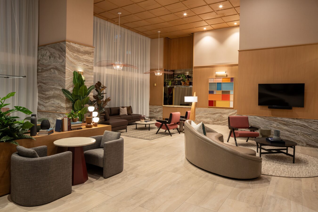 Modern hotel lobby with stylish seating areas, plants, a wall-mounted TV, colorful artwork, round tables with chairs, and warm lighting. Marble and wood accents create a cozy, inviting atmosphere.