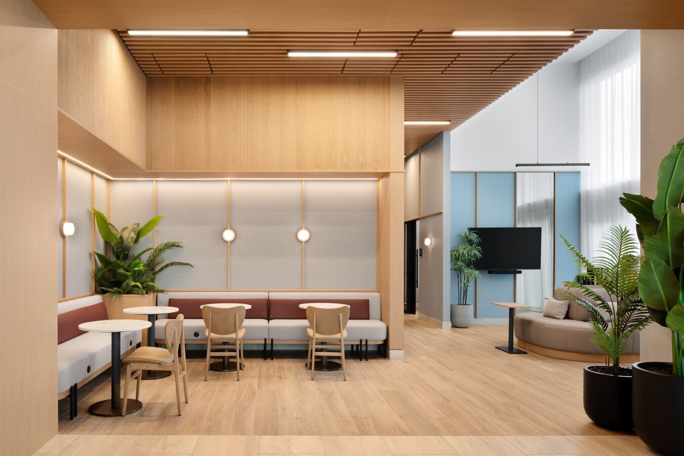 A modern, spacious lounge with light wood floors and walls, booth and chair seating, round tables, large green plants, and soft lighting. There is a sofa and a TV in the background, with tall windows letting in natural light.