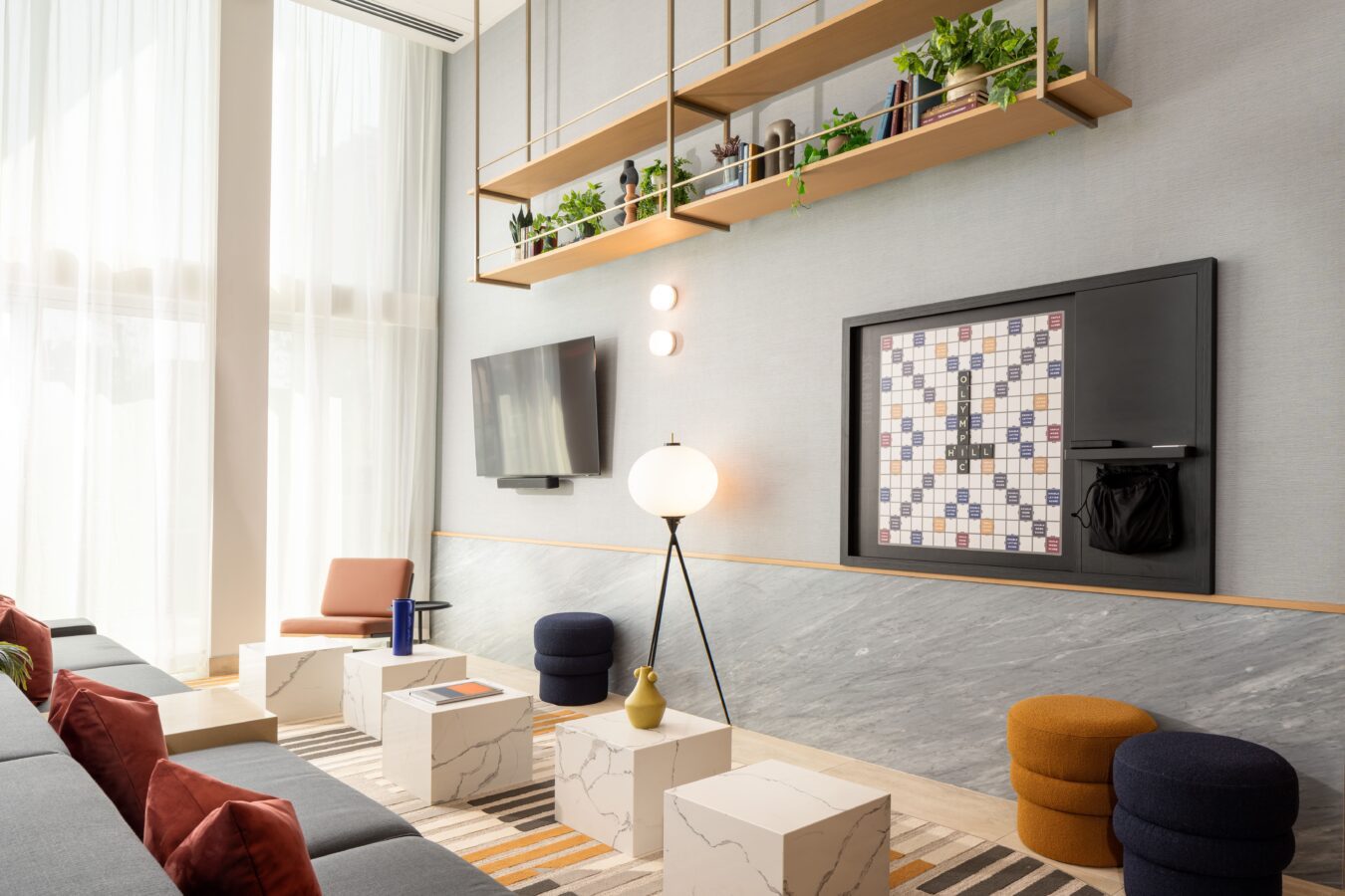 Modern lounge with marble cube tables, colorful poufs, a striped rug, a wall-mounted Scrabble board, a TV, tall windows with sheer curtains, a floor lamp, and shelves with plants above.
