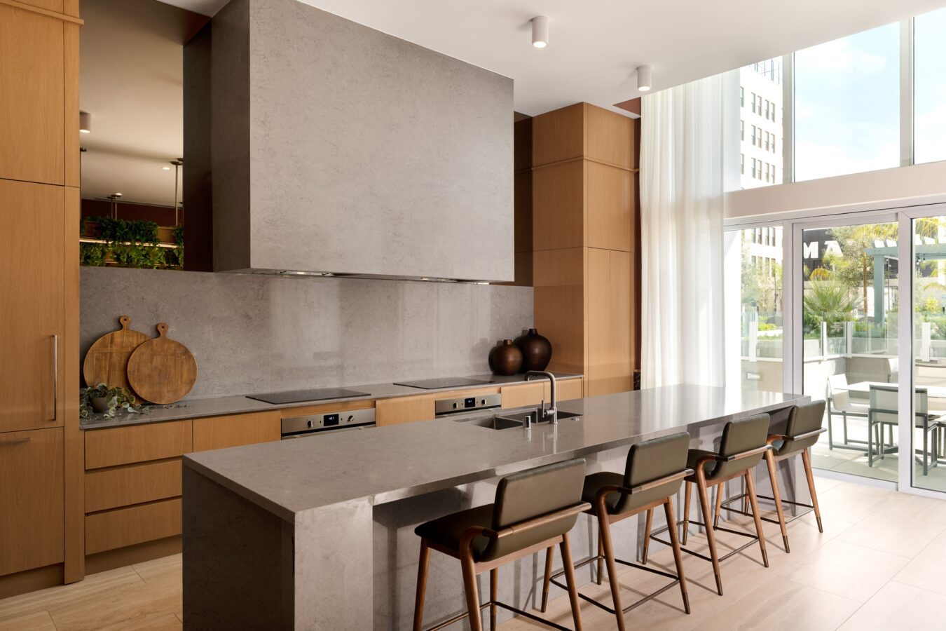 Modern kitchen with light wood cabinets, a long gray stone island with sink, four bar stools, stacked ovens, and large windows letting in natural light. Cutting boards and decorative vases are on the counter.