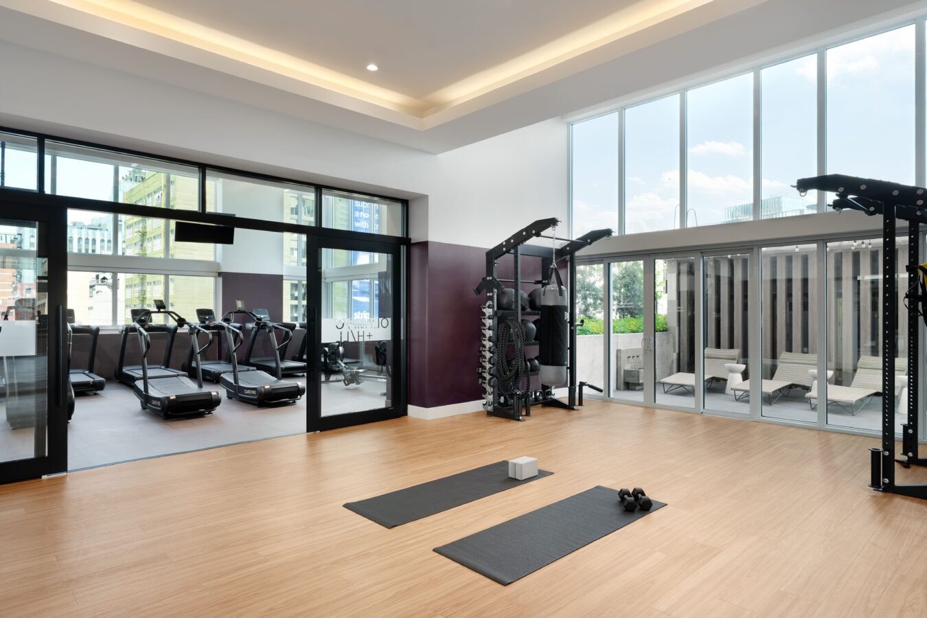 A modern gym with large windows, wooden floors, and two yoga mats with blocks and dumbbells. In the background, treadmills are visible through glass doors, and outdoor seating can be seen through more windows.