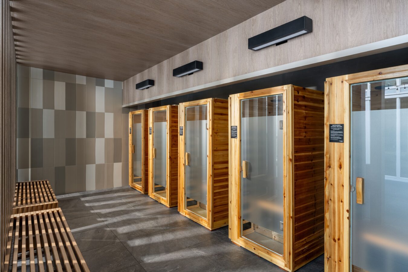 A modern wellness facility with four wooden sauna cabins featuring frosted glass doors, set against a tiled wall. Wooden benches line the opposite wall on a dark tiled floor.