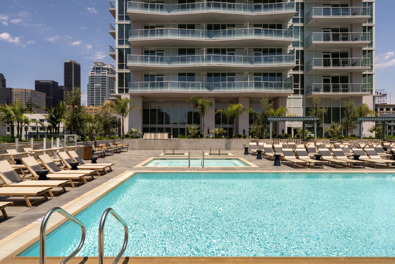 A modern apartment building with balconies overlooks a swimming pool area with lounge chairs and palm trees; a city skyline is visible in the background under a clear sky.