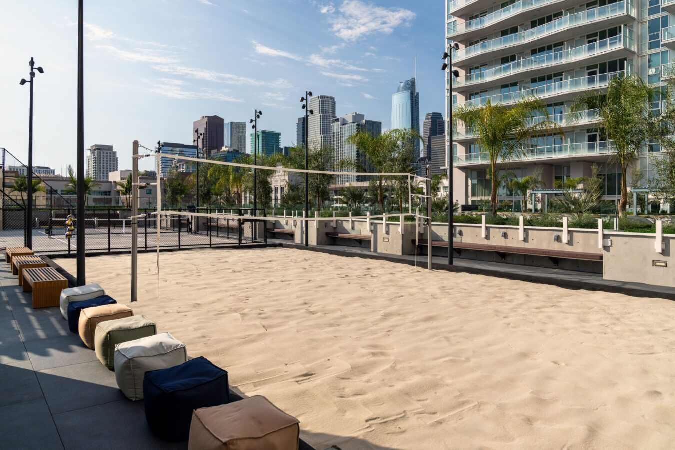 A sandy volleyball court with a net is set outdoors, surrounded by benches and colorful cushions, with tall modern buildings and palm trees in the background under a partly cloudy sky.