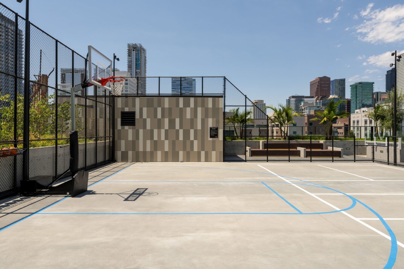 A modern outdoor basketball court with blue and white lines, a hoop, and a fenced area, set against a backdrop of tall city buildings and palm trees on a sunny day.