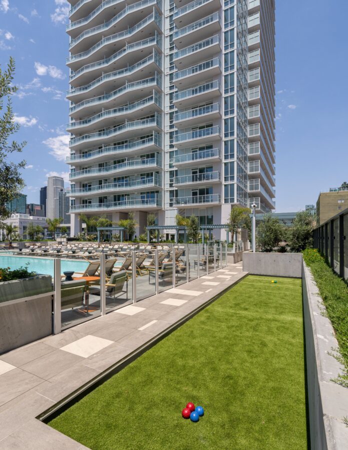 A modern high-rise apartment building with balconies overlooks a pool area with lounge chairs and palm trees. In the foreground is a bocce ball court with red and blue balls on green turf. The sky is clear and sunny.