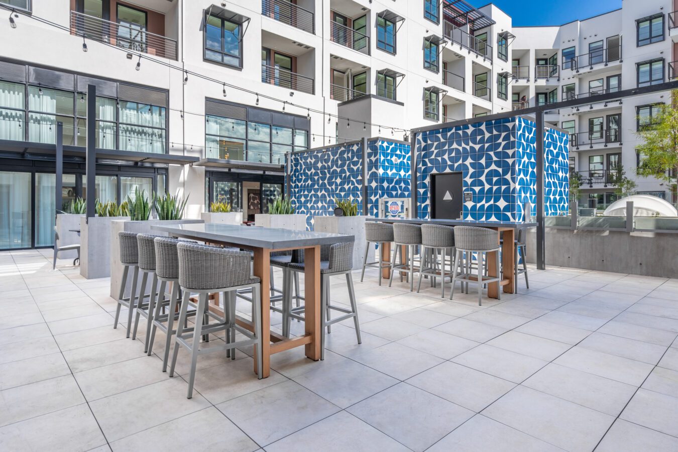 Modern outdoor patio with tall tables and woven barstool seating, set against a white apartment building. Blue geometric patterned walls provide accent, and large windows overlook the area. The space is clean and bright.