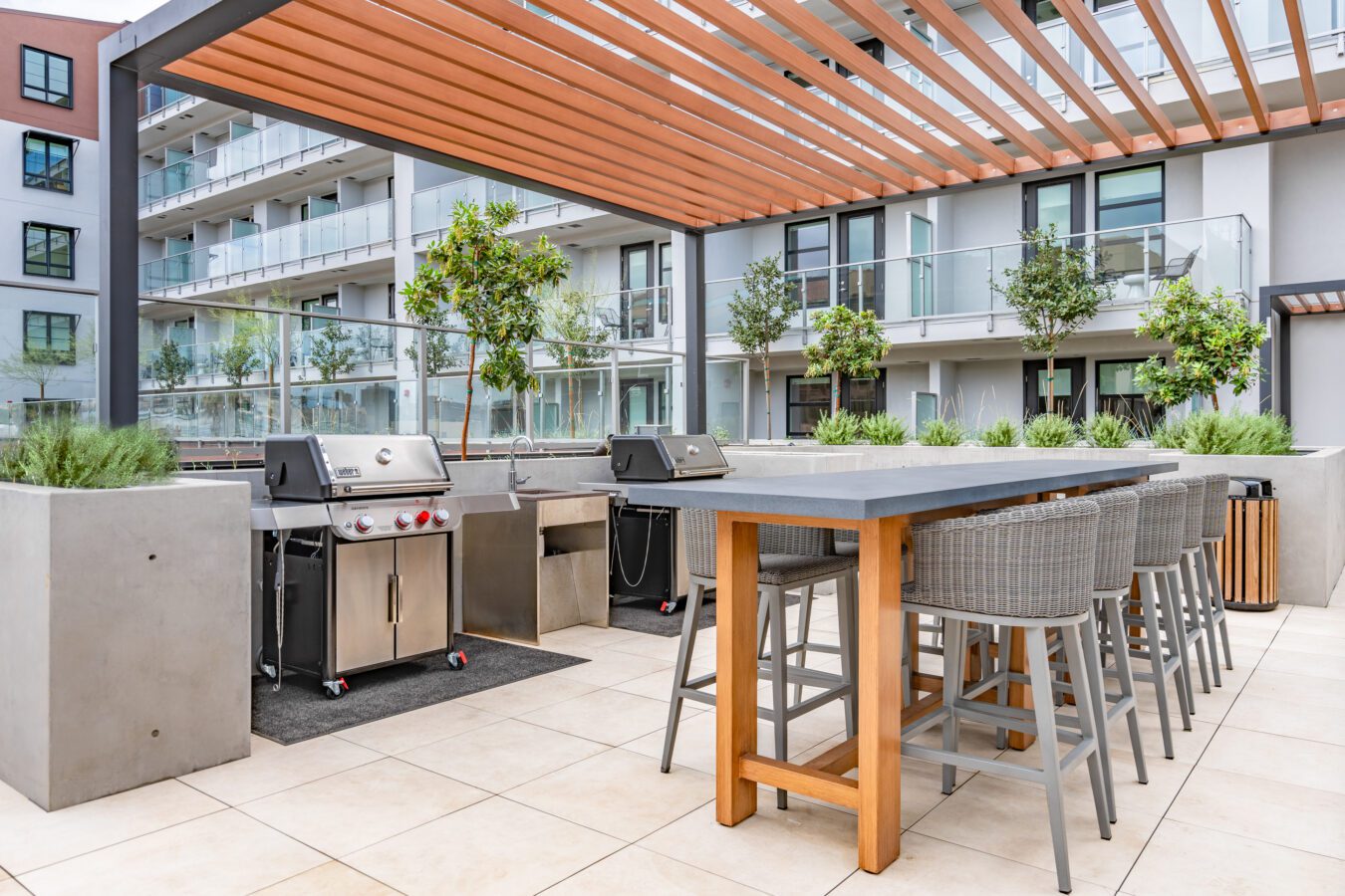 Modern outdoor patio area with two stainless steel grills, a long dining table with wicker chairs, planters with greenery, and a wooden slatted pergola, set against a backdrop of apartment buildings and balconies.