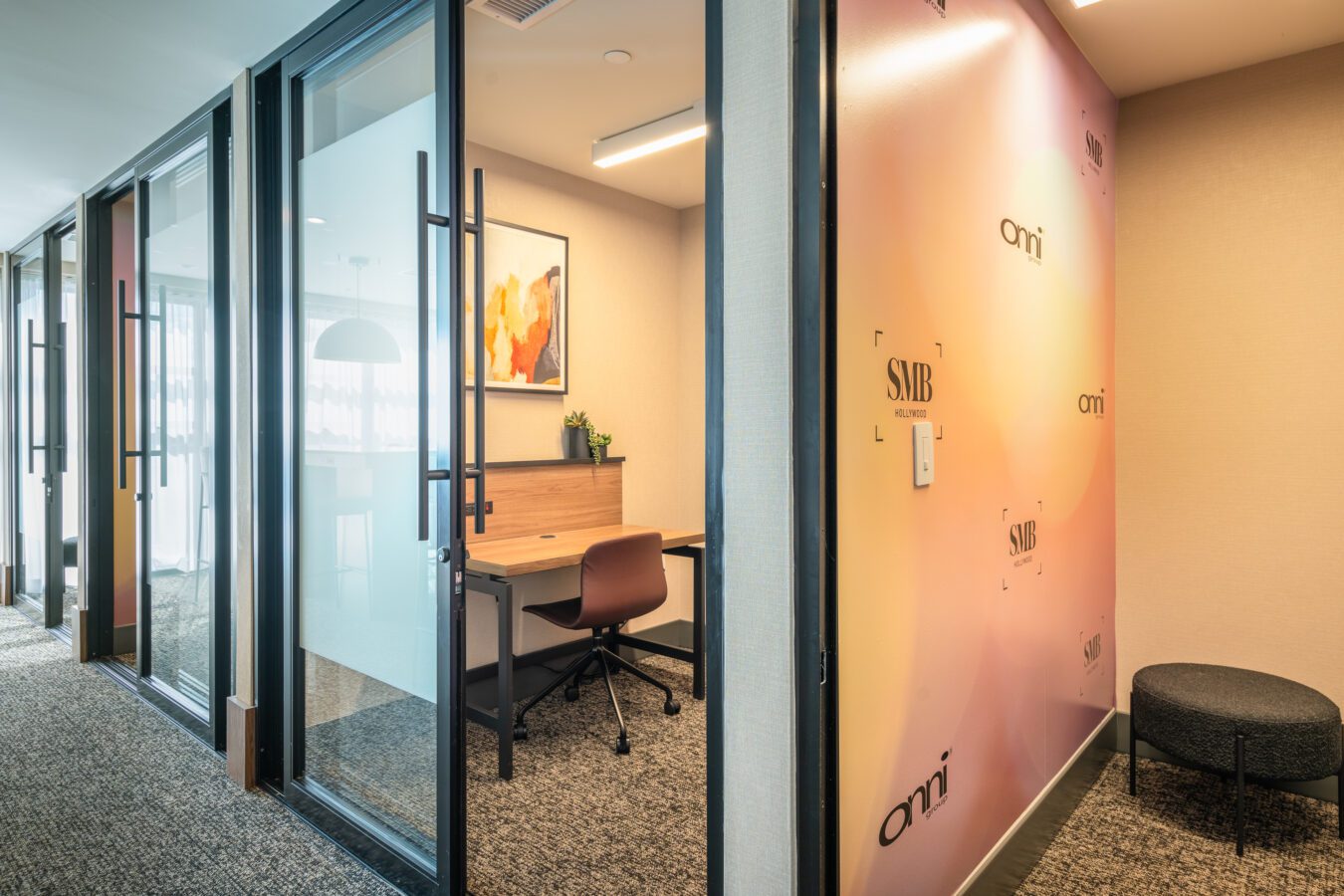 Modern office hallway with glass-walled cubicles, each with a desk and chair. A colorful wall with the text SMB and ami is on the right next to a small seating area with a round ottoman.