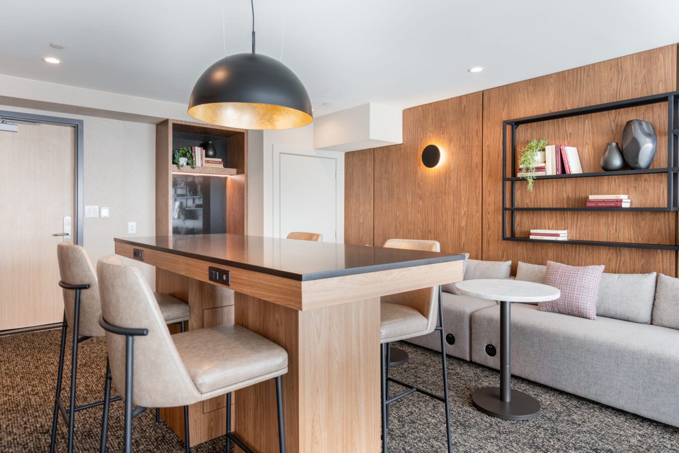 Modern lounge area with a high wooden table, four beige chairs, a grey sofa, a small round table, wall shelves with books and vases, and a large black hanging light fixture. The decor is minimalist and contemporary.