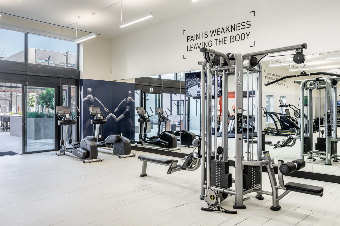 Modern gym with cardio and weight machines, a bench, large mirrors, and wall art of running figures. A quote on the wall reads “Pain is weakness leaving the body.” Bright and clean with large windows.
