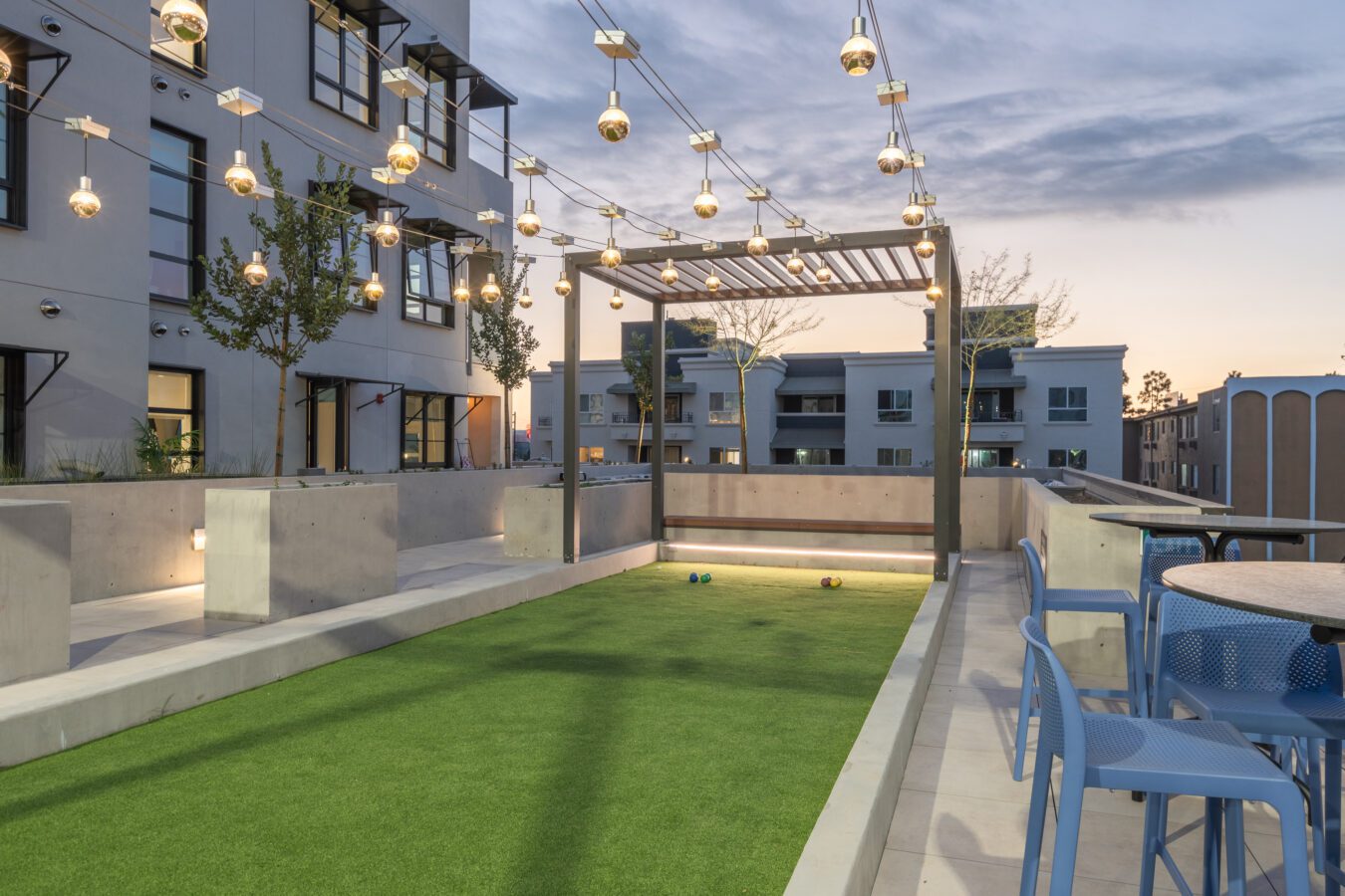 A modern apartment rooftop with string lights, a pergola, and a bocce ball court. There are concrete benches, outdoor tables and chairs, and a view of surrounding buildings at sunset.