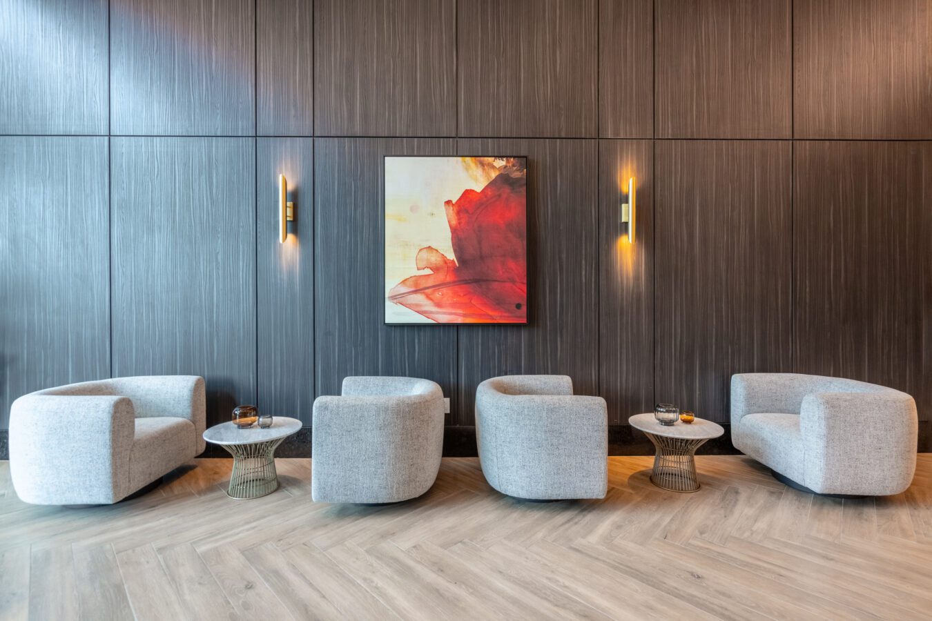 A modern lounge area with four light gray armchairs, two round side tables, and a red abstract painting on a dark wood-paneled wall, illuminated by two wall sconces.