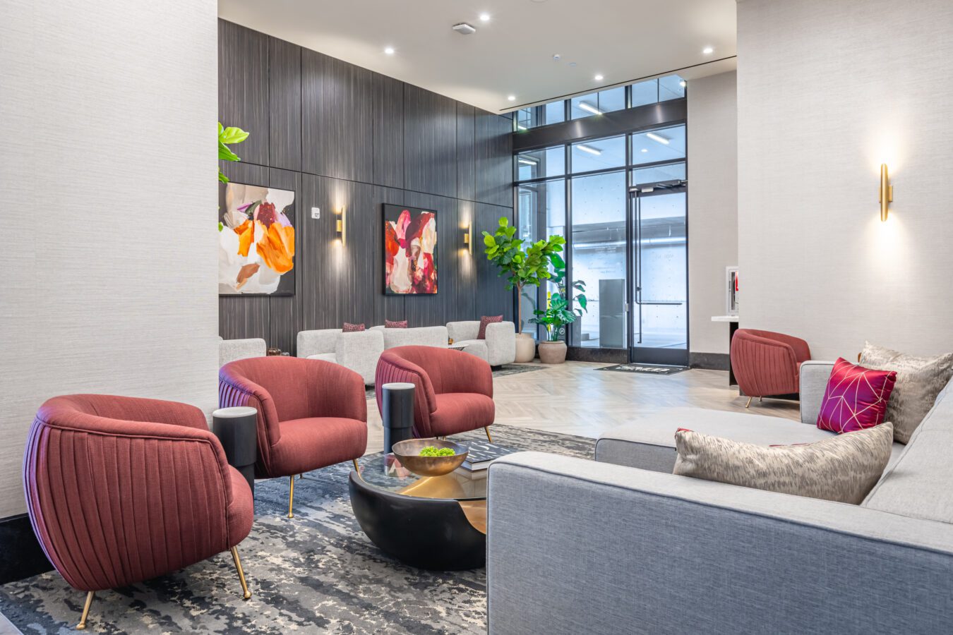Modern lobby with gray sofas, red armchairs, abstract art on the walls, large windows, potted plants, and a central black coffee table with a bowl. The space is well-lit and contemporary in design.