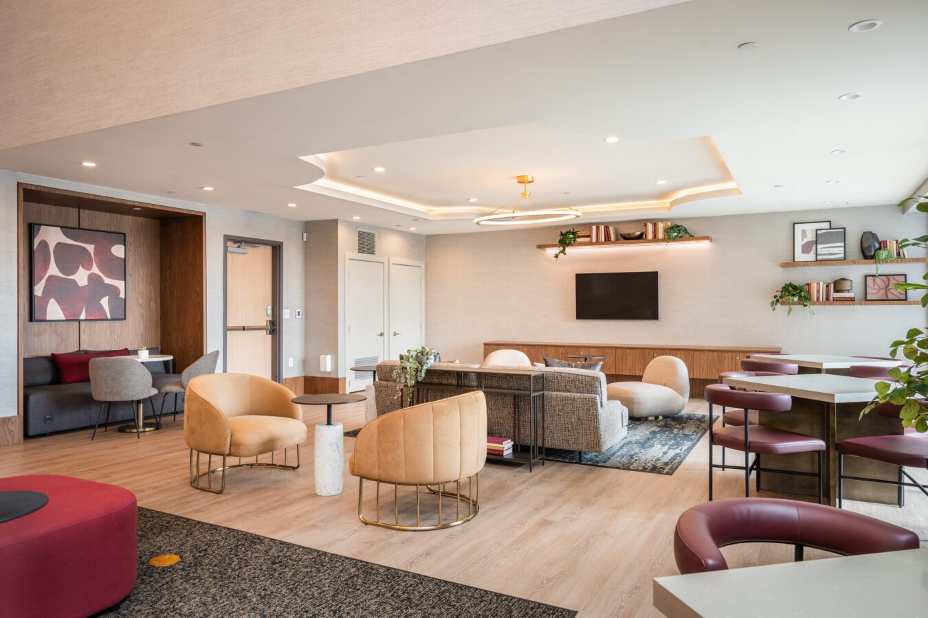 Modern lounge with beige walls, contemporary furniture including cushioned chairs, sofas, and tables. There’s a TV on the wall, plants, shelves with decor, and a large window letting in natural light.