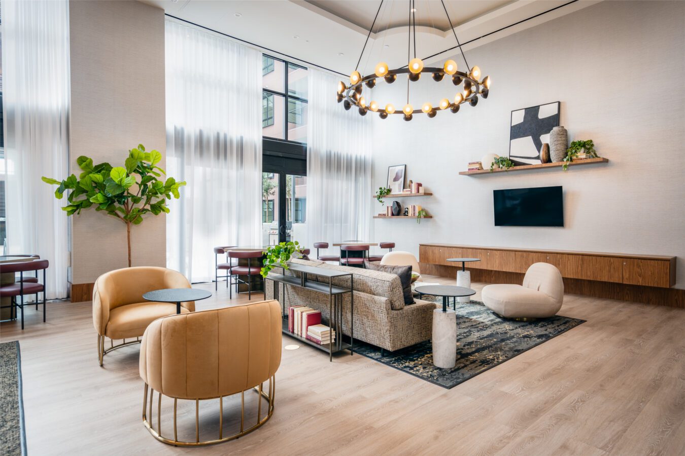 Modern lounge with high ceilings, large windows, neutral-toned furniture, round chandelier, indoor plant, and wall-mounted shelves with decor. Tables and chairs are arranged for casual seating and conversation.