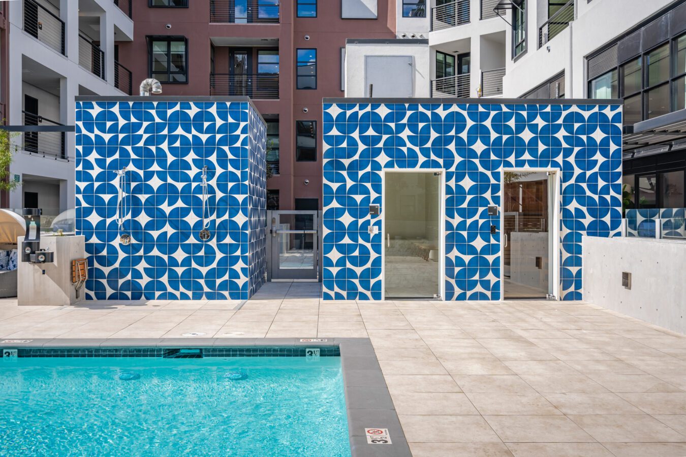 A modern outdoor pool area with two adjacent buildings covered in blue and white geometric tile patterns, featuring open doorways and surrounded by apartment buildings.