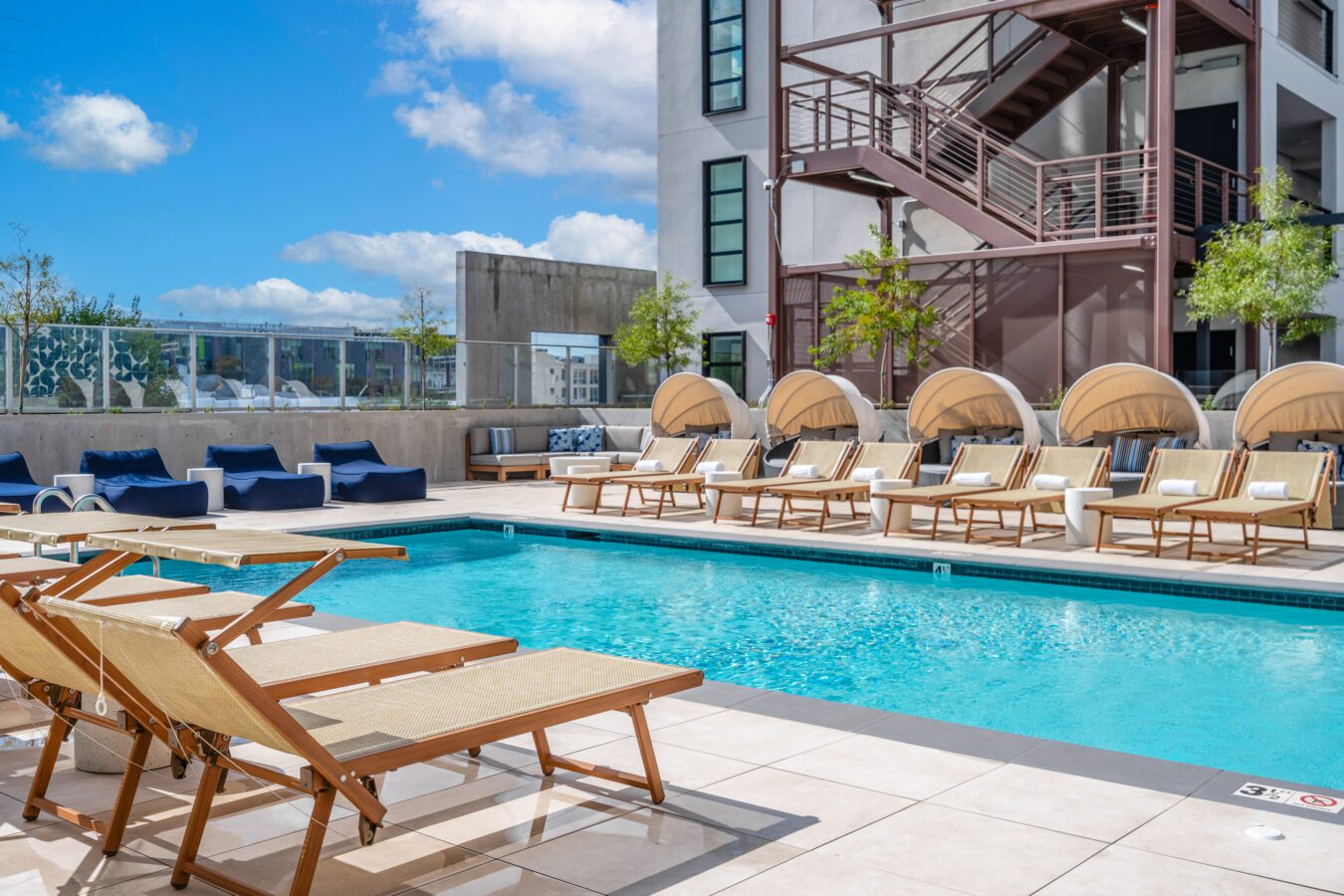 A modern outdoor swimming pool with wooden lounge chairs, round cabanas, and blue cushioned seating, surrounded by a building with exterior stairs under a bright blue sky with clouds.