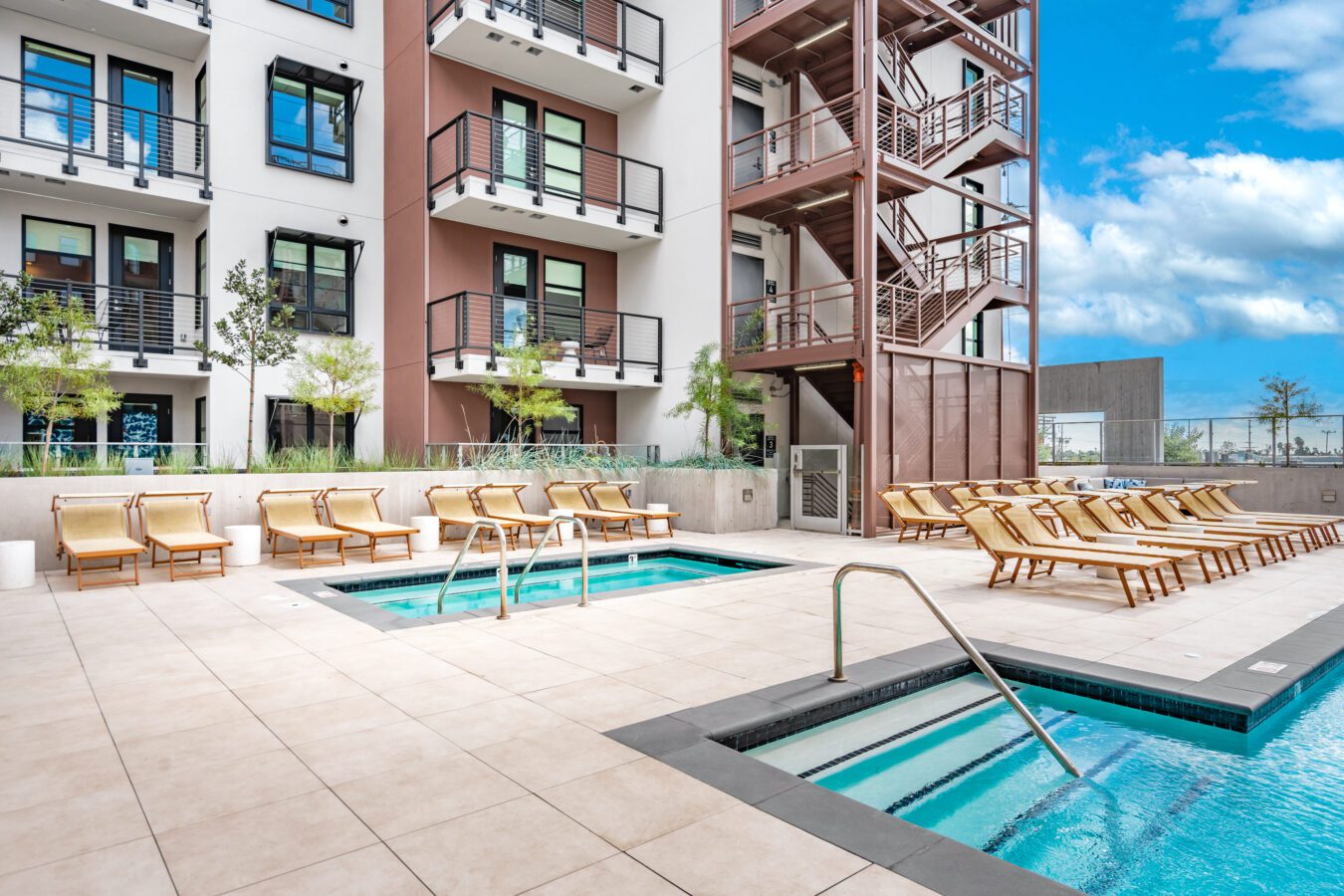 Modern apartment building with balconies overlooks a clean outdoor pool area, featuring two small pools or hot tubs surrounded by tan lounge chairs on a spacious tiled deck under a blue sky with clouds.