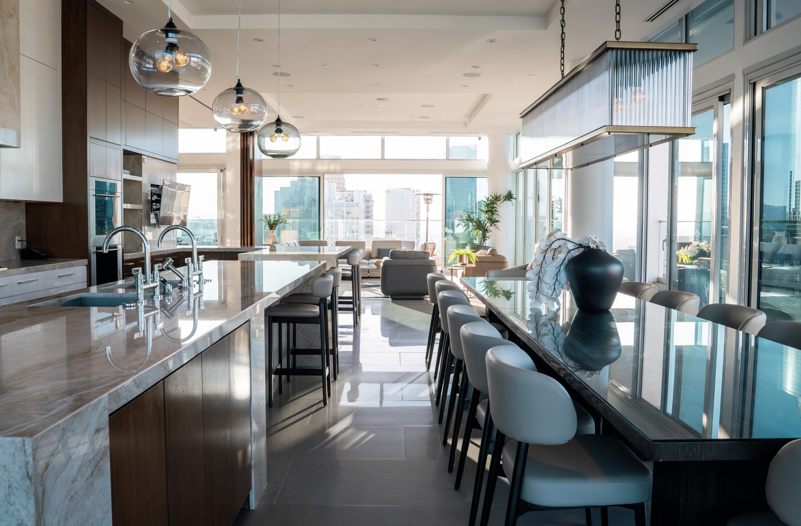 Modern, spacious kitchen and dining area with large island, marble countertops, contemporary pendant lighting, bar stools, and floor-to-ceiling windows offering a city skyline view, with sunlight streaming in.