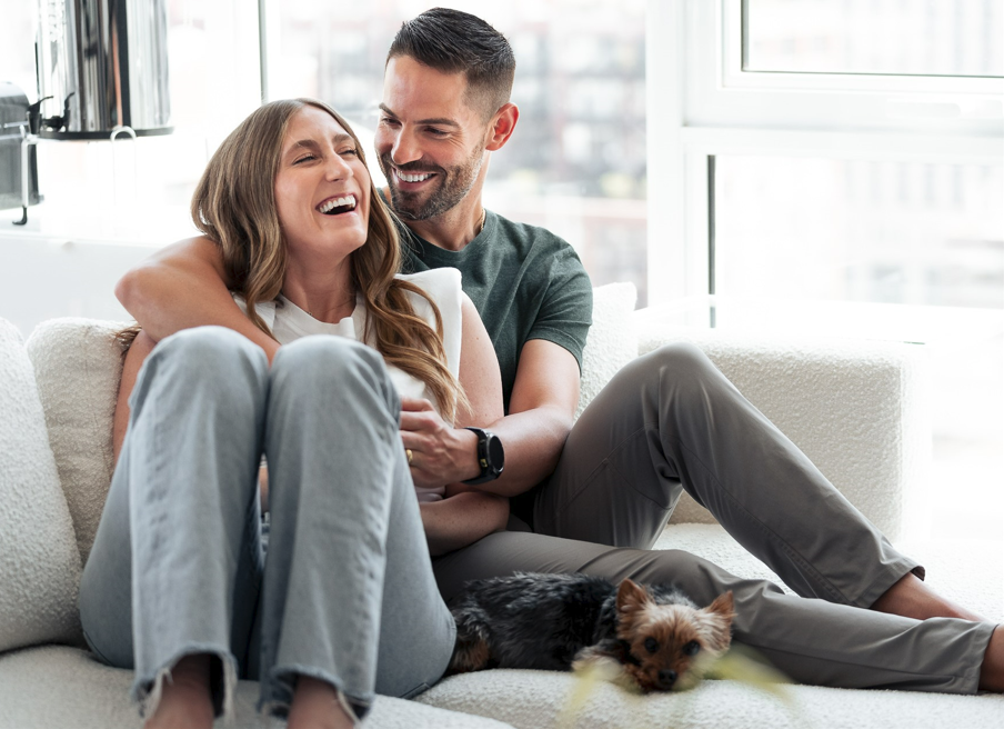 couple sitting on couch laughing together with their little dog