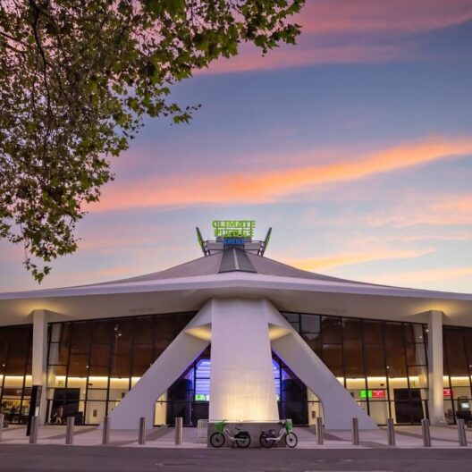 A modern arena with angular white pillars and large glass windows is shown at sunset. A neon sign on top reads Climate Pledge Arena. Bicycles are parked in front, and pink clouds fill the sky.