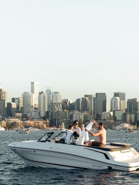 Four people enjoy a sunny day on a white speedboat in the water, with a city skyline of tall buildings in the background. The group appears relaxed and is socializing, with clear skies overhead.